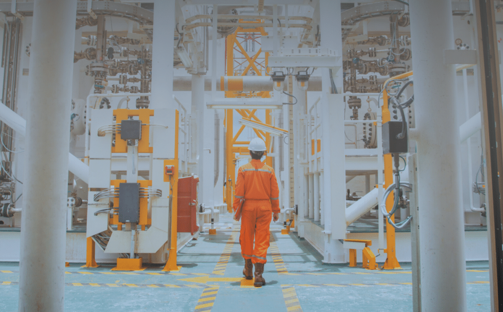 A person wearing an orange jumpsuit and a white hard hat walks through an industrial facility filled with machinery and pipes.