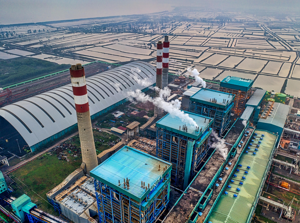 An aerial view of an industrial facility with multiple large chimneys emitting white smoke, surrounded by vast, organized rows of agricultural fields. The image captures the intersection of industrial activity and expansive land use, showcasing the contrast between the facility's blue structures and the green landscape.