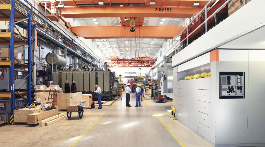 A spacious industrial manufacturing facility with high ceilings, overhead cranes, and large electrical transformers in various stages of assembly. Several workers wearing safety helmets and uniforms are engaged in discussions and tasks, while wooden crates and equipment are organized throughout the space. A modern control panel is mounted on the wall, indicating a well-structured and automated production environment.