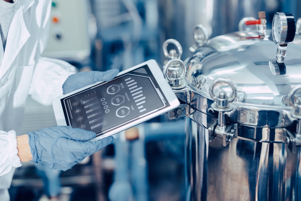 The image shows a person wearing blue gloves and a white lab coat, holding a tablet near a large stainless steel industrial vessel or reactor. The tablet screen displays various metrics, including water level, pressure, and temperature, with graphical bars and percentage indicators. The setting appears to be a laboratory or industrial facility, likely focused on monitoring and controlling processes within the vessel using the digital interface on the tablet.